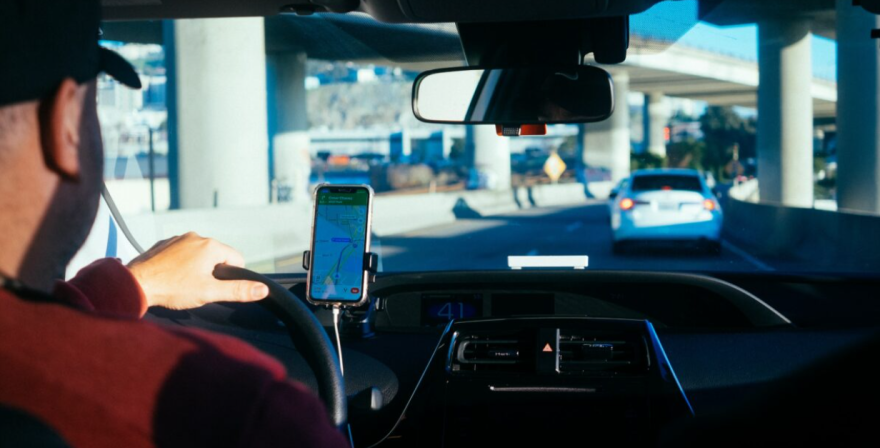 A man with light white skin tone drives a car. Only part of his side profile is seen, he has on a red long-sleeve. The focus is on a phone displaying GPS directions.