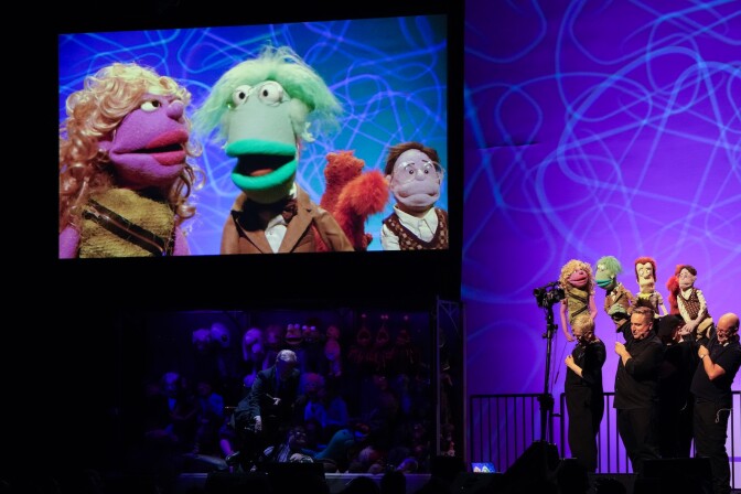 A view of about three puppeteers dressed in black on a stage with a large screen to their left showing a purple female puppet a green male one, a purple male one with glasses in the back and what appears to be a squirrel puppet next to him.