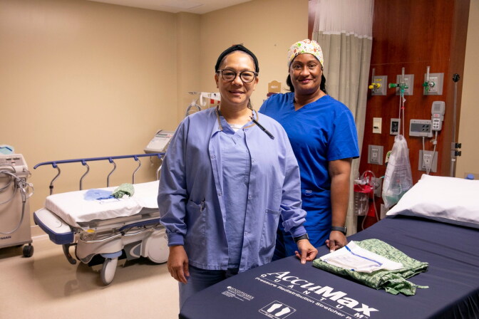 Two people stand in scrubs at the side of a hospital bed, another is in the background of the hospital room.