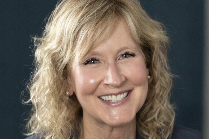 A woman with blonde, shoulder length hair, smiles while seated in front of a black background wearing a black blazer 