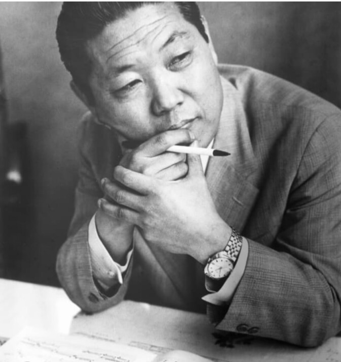 A black and white photo of a middle-aged Korean man in a suit staring off pensively to the side, while holding his chin in one hand. 