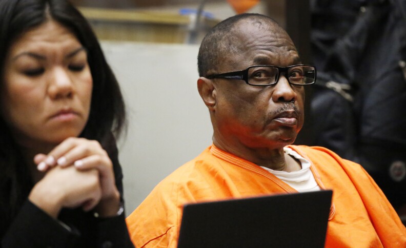 A man with dark-tone skin and glasses sits in an orange jumpsuit.