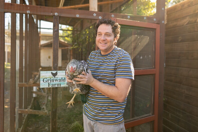 A white man with brown hair wearing a blue, white, and yellow striped t-shrit, stands next to a wooden chicken coop holding a black and white spotted chicken.
