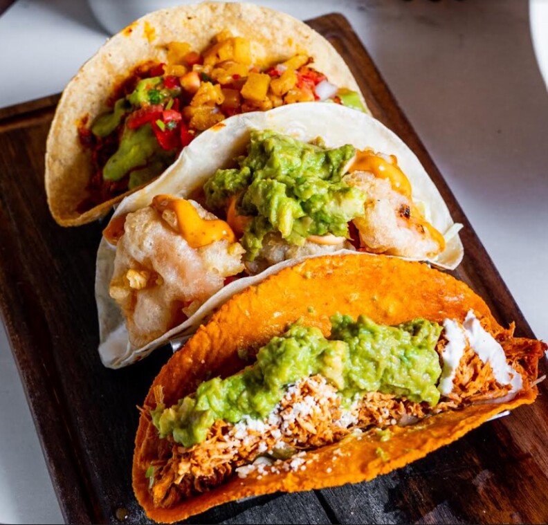 An image of three tacos with different colored tortillas on a dark wood plank