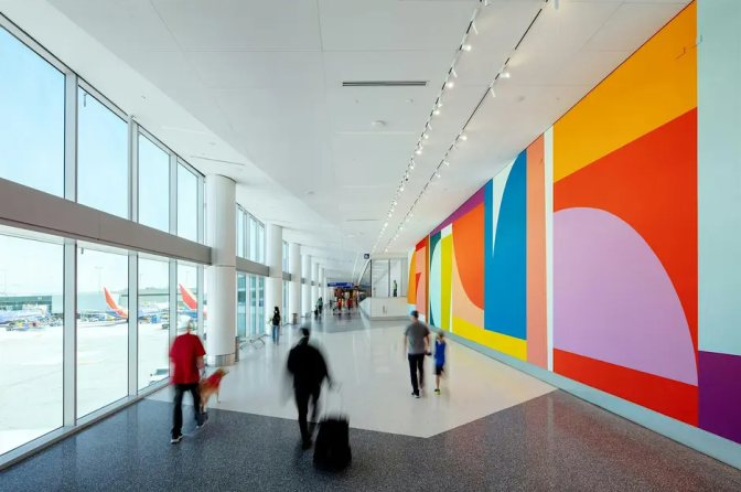 Travelers walk through an airport terminal past a colorful geometric mural and floor-to-ceiling windows overlooking the tarmac.