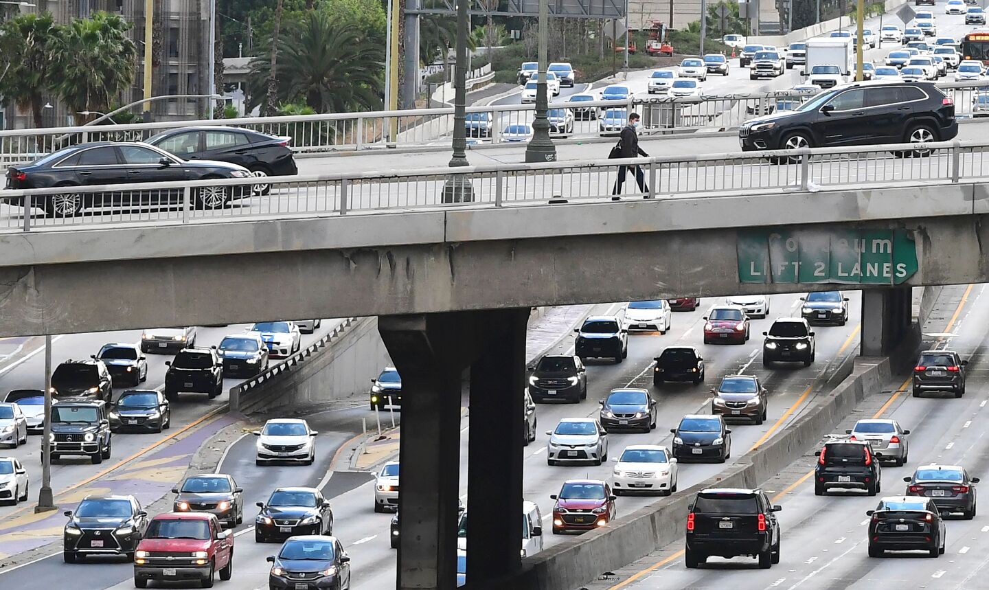 Cars crawl along the 110 Freeway through downtown Los Angeles as a person walks across on an overpass above.
