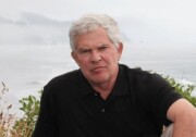 A white man with silver hair wearing a black polo shirt sits in front of a lake