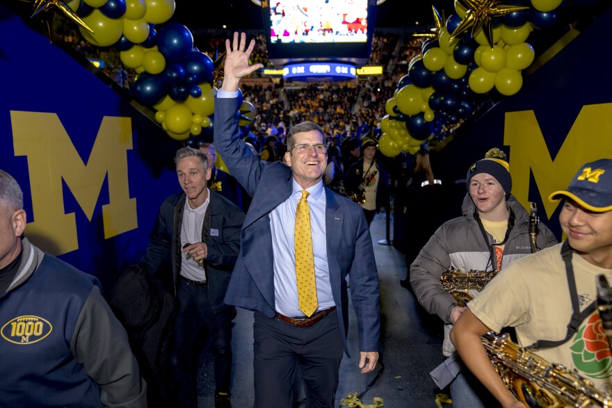A white man in a suit walks through an alley at a stadium, and waves to fans decked out in maize and blue colors.