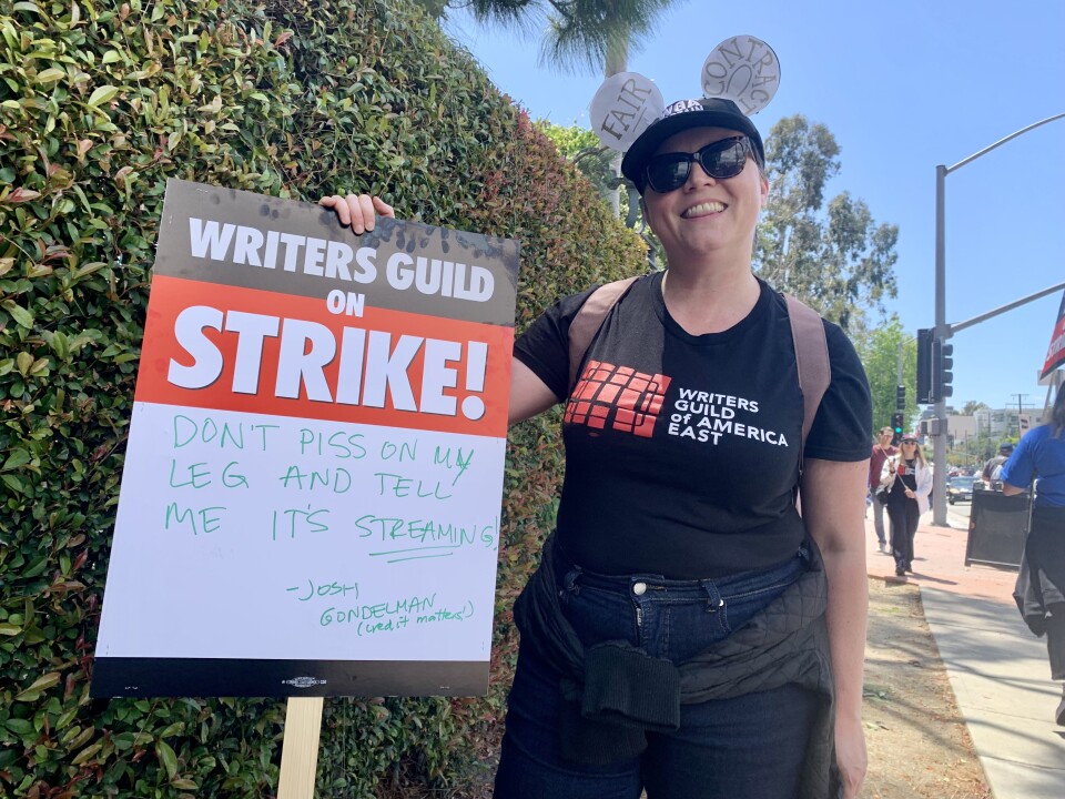 A woman with short hair under a black cap holds a sign that reads "Writers Guild on STRIKE!"  Handwritten is "Don't Piss On My Leg And Tell Me It's Streaming." She wore Mickey Mouse ears that read "Fair Contract." 