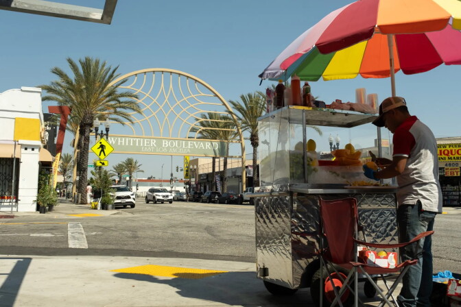 A street vendor stands under a rainbow umbrella peeling fruit in front of a cart. In the distance is an arch with a sign that reads "Whittier Blvd. East Los Angeles."