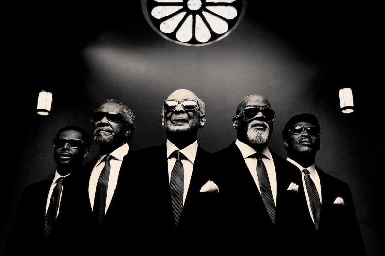 Five Black men stand in dark suits and dark glasses in this black-and-white image.