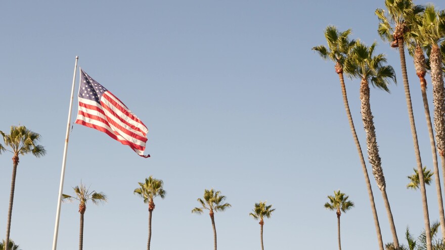A U.S flag flies against a clear gray/blue sky with a row of tall palms.