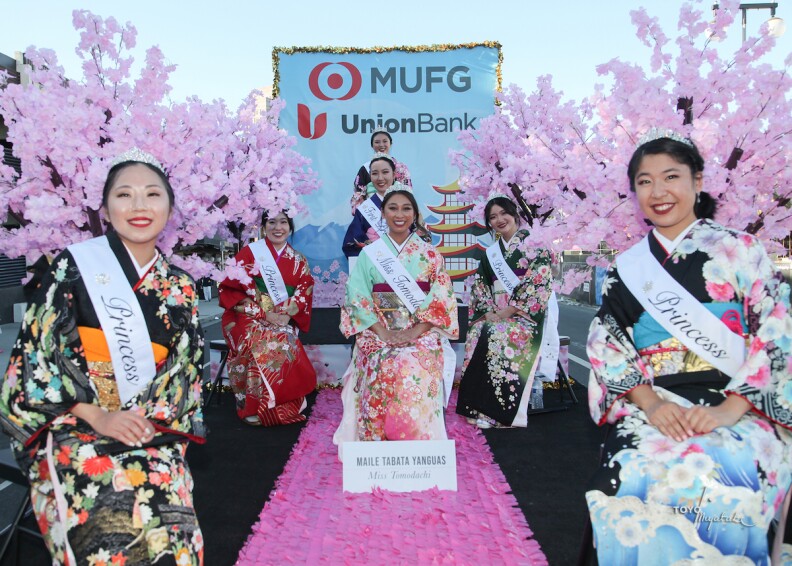 The Nisei Week queen sits on a pink carpet while her court of six flanks her. They are all wearing sashes and wear kimonos,