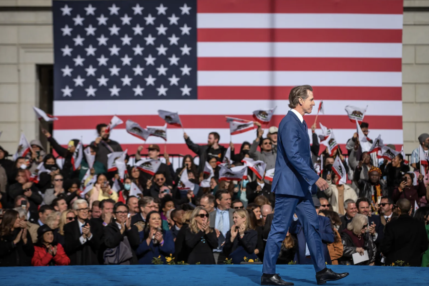 A man with light skin tone wearing a suit walks left to right across a stage. A large crowd and an American flag is in the background.