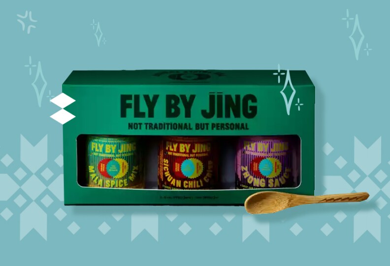 A green box containing three small glass jars all with different colored labels, a wooden spoon sits outside the box.