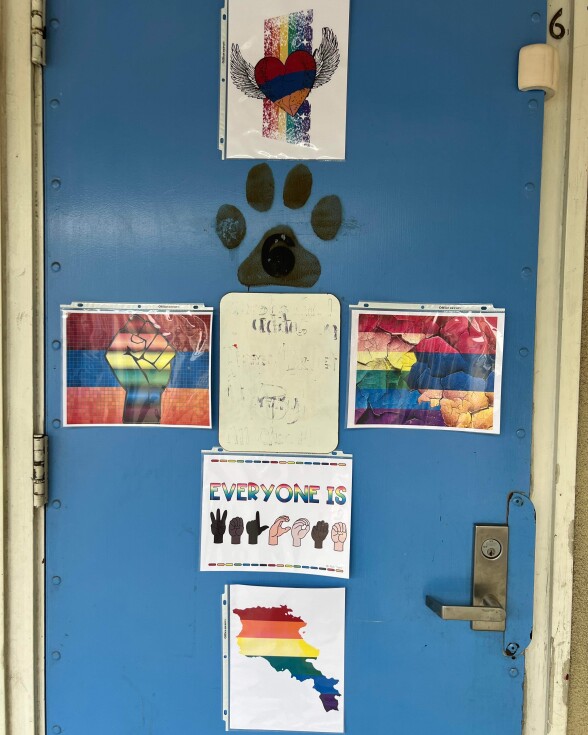 A view of the teacher's blue door with multiple versions of Pride and Armenian flags printed on paper in protective sleeves. On the left is a rainbow illustrated fist in front of the Armenian flag colors. On the right is both flags crossing into each other with a rocky texture.