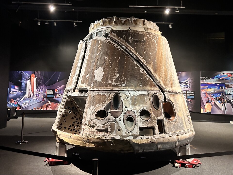 The dirty and charred backside of a large spacecraft on display in a dark, large room. Several colorful rendered images are displayed on the wall behind it.
