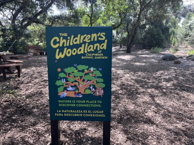 A big green sign in the middle of a forested area says "The Children's Woodland at California Botanic Garden." It also bears an illustration of a tree.