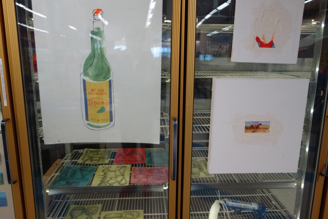 A painting of a hot sauce bottle and several other paintings and sketches in a repurposed freezer.
