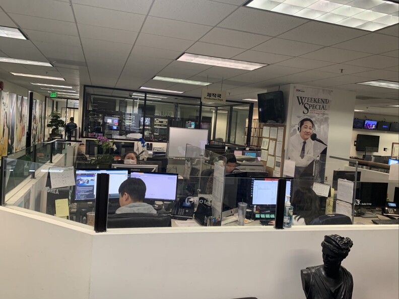 Two women and a man sit at computer terminals inside a radio station newsroom as another person in the distance walks down the hallway.