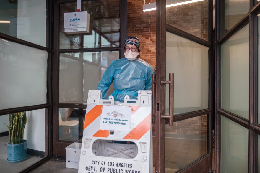 A woman dressed in full protective gear including a face mask places an orange and white barrier in front of an open door.