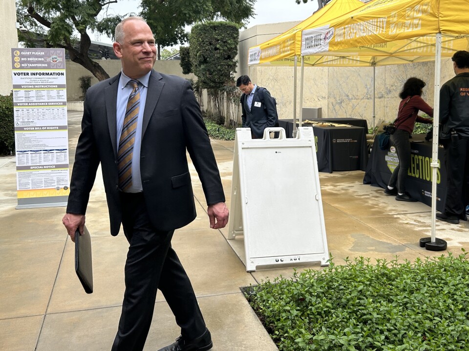 A man in a suit smiles at something off camera as he walks in front of a yellow tent with people working underneath. 
