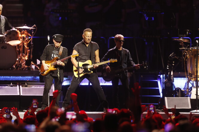 A man who appears to be white and in his 60s or 70s performs onstage with a guitar. Two other men playing guitar flank him. A large crowd with their hands in the air can be seen in front of the stage. 