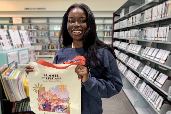 A woman with dark skin tone and long dark brown hair smiles and holds up a cream colored canvas totebag that reads Summer with the Library and has a colorful scene of Los Angeles including a palm tree, sun, and prickly pear cactus. She is surrounded by full bookshelves. 