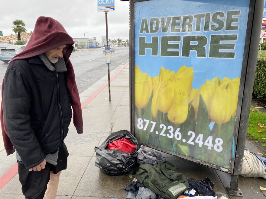 A man in a black jacket with a red hoodie over his head looks down at piles of clothing and a soaked red sleeping bag on the ground next to a road. A bus station advertising board is in the background. 