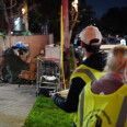 Two people wearing reflective vests stand next to a makeshift shelter on the sidewalk.