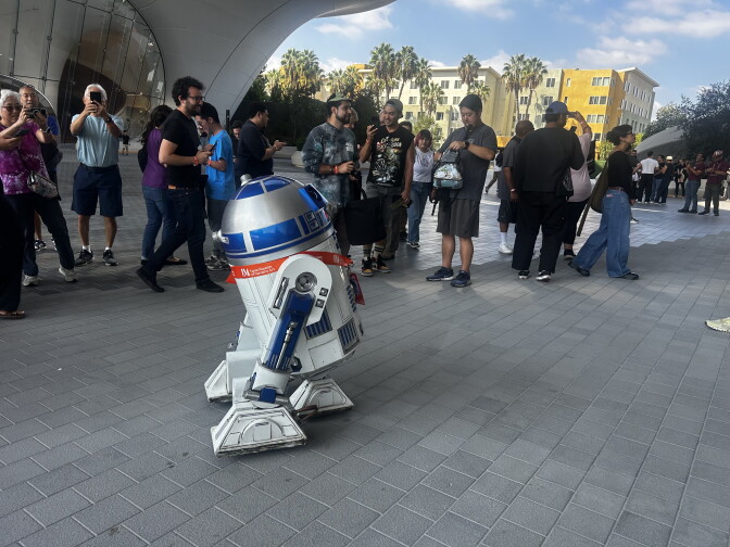 A life-size R2-D2 droid rolls through a plaza as a crowd takes photos on their phones outside a building.
