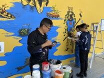 Three people work on a mural, which features a body of water and other elements, including a fisherman casting a net and a lowrider sprouting flowers. There are painting tools around the three people, including a ladder and multiple buckets of paint. The floor has been sealed off with plastic and blue tape. 