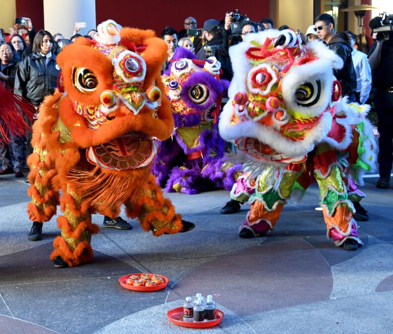 People in an orange Chinese dragon costume on the left, others in a white Chinese dragon costume on the right, with a purple dragon behind in front of a crowd of onlookers. They stand on outdoor tile, small plates with other items in front of them.