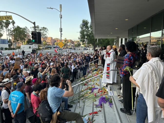 A group of people of varying ages, genders and skin tones stand at the bottom of concrete steps. A man wearing white robes stands at the top of the steps, which are covered in flowers. 