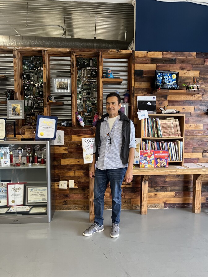 A dark-haired man with medium skin stands in a community resource center. He's wearing jeans and a blue button up shirt with a sweater hanging on his shoulders.