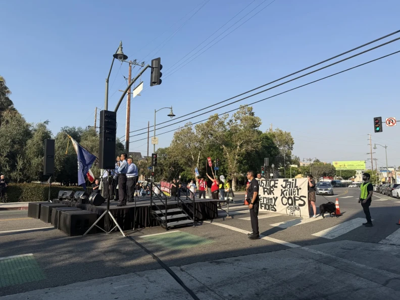 Protesters holding signs and banners during a demonstration on a street, with speakers standing on a stage and police officers monitoring the event.