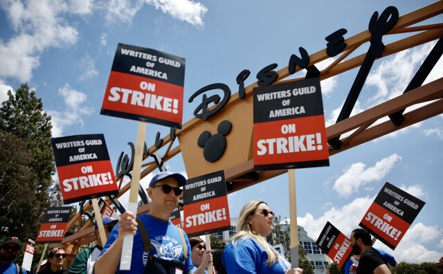 A group of people wearing blue T-shirts and holding black and white signs with the words "Writers Guild of America on Strike!" are picketing underneath a big sign that reads "Walt Disney." Scattered wisps of clouds break up the otherwise blue sky on a sunny day, and the man and woman closest to the viewer are wearing sunglasses. 