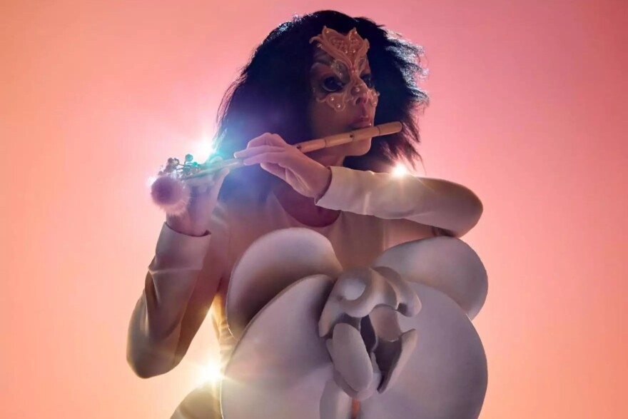 Björk, lit from behind and wearing a gold dress, with a golden tiara, and a series of white shapes in front of her, plays a flute.
