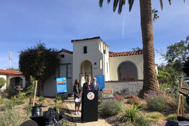 A wide shot of a man speaking at a black podium in front of an adobe-style home. A large palm tree is in the background and native plants are in the front yard. 