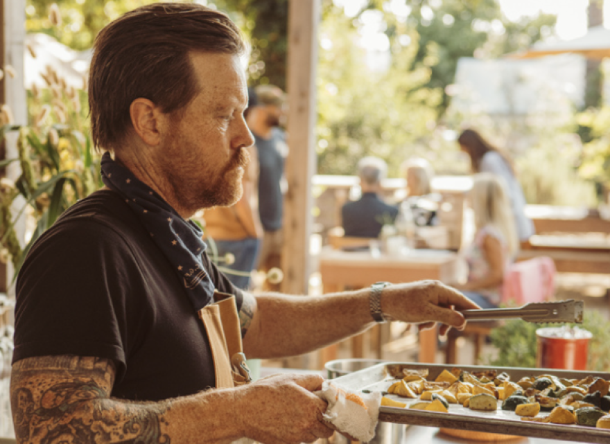A light skinned man with reddish hair and beard and tattos on his arm is wearing a black T shirt and a blue dotted bandana. He's looking at a tray of cooked potatoes with silver tongs in his hand.