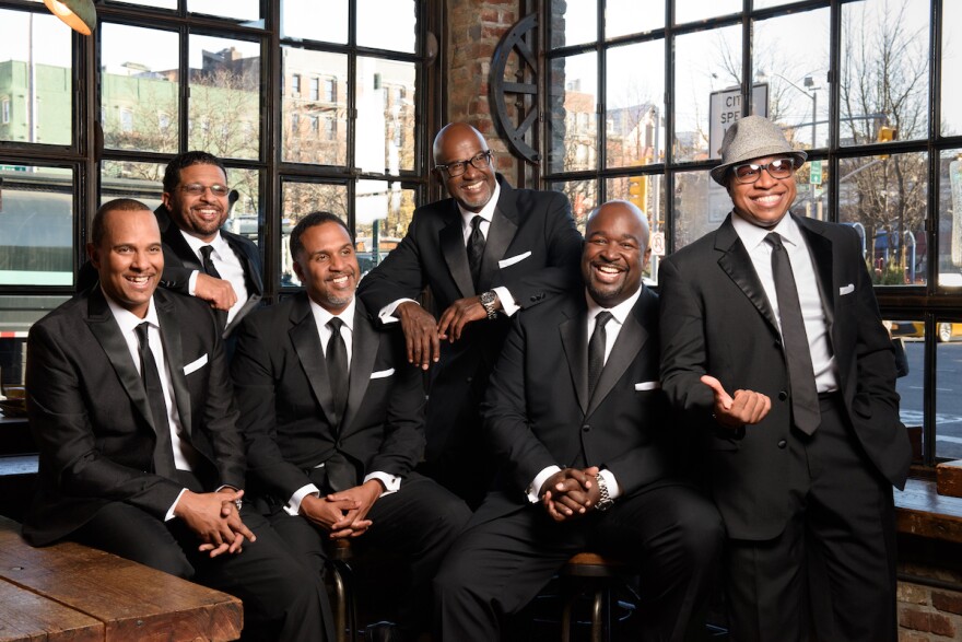 The six African American men who comprise the vocal group Take 6 wear black suits as they sit or stand next two walls of windows