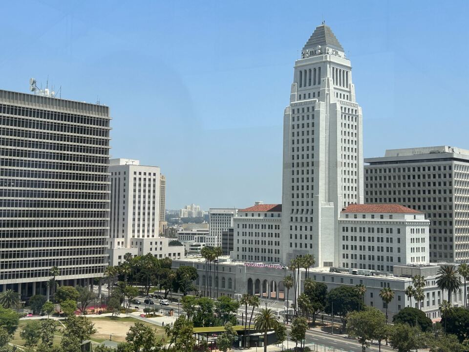 A tall, white building is surrounded by shorter buildings and trees during the day.