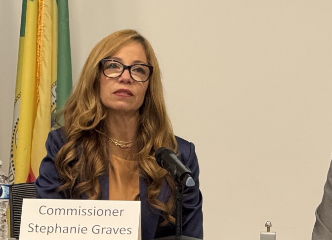 A woman with glasses and a dark blue suit jacket looks forward in front of a microphone and a sign saying "Commissioner Stephanie Graves."