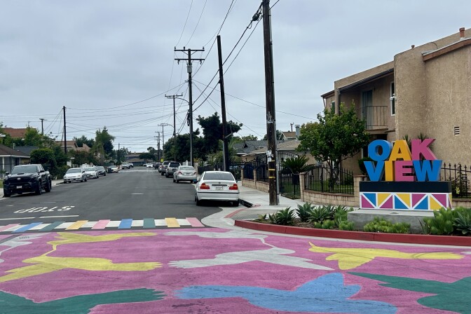 At an intersection in a residential neighborhood, a colorful sign reads "Oak View" and there is a pink, white, yellow, blue, and green pattern painted on the asphalt across the intersection.