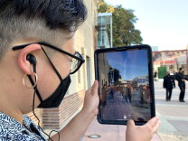 The side profile is visible of a person with short hair and hoop earrings, holding a tablet with an image of people in 1940s-era dress superimposed over the present-day scene of a courtyard.