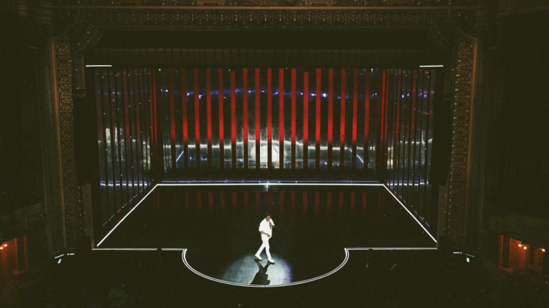 This is a wider shot of a black stage with white trim. On it stands one man with dark skin tone and dressed in white, speaking to an audience through a microphone.