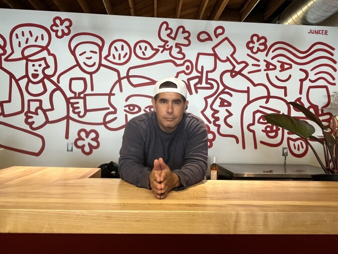 A light-medium toned man leans over a wooden counter with a background of a mural in white and red figure drawings and a plant to the right.