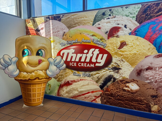 A life-sized cardboard cutout of Thrifty Ice Cream's smiling cone mascot giving two thumbs up, standing in front of a wall mural of colorful ice cream scoops and the Thrifty Ice Cream logo, "Since 1940."
