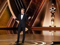 Conan O'Brien stands on the Oscars stage wearing a black tux with a bow tie. His arms are outstretched at the elbows as he speaks to the audience. A tall golden Oscar statue is behind him on stage.