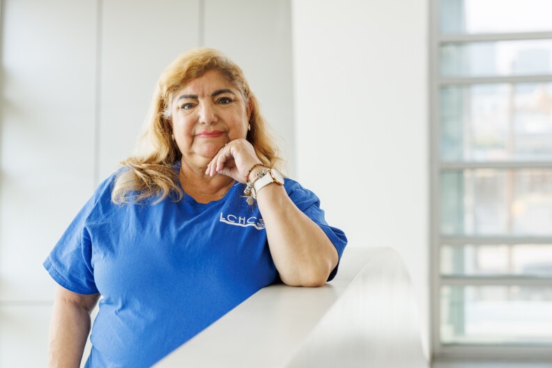 A woman wearing a blue tshirt rests her chin on her hand as she leans against a white wall.